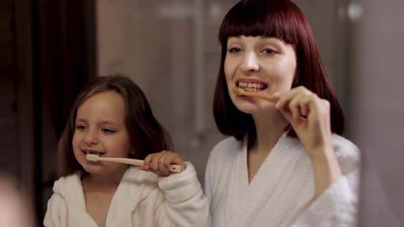 Mom and Her Cute Girl in White Bathrobe Brushing Teeth Together Standing Near the Mirror