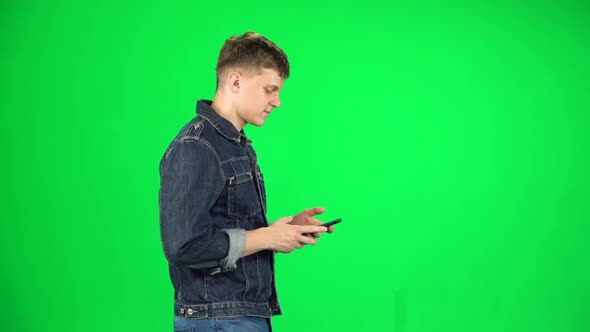 Smiling Man Goes and Texting with Smartphone on Green Screen at Studio. Slow Motion. Side View
