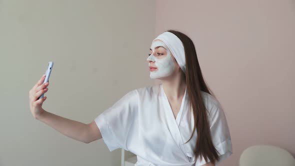 Young Woman in Beauty Cosmetology Salon with Clay Mask on Face Uses Phone to Takes a Photo