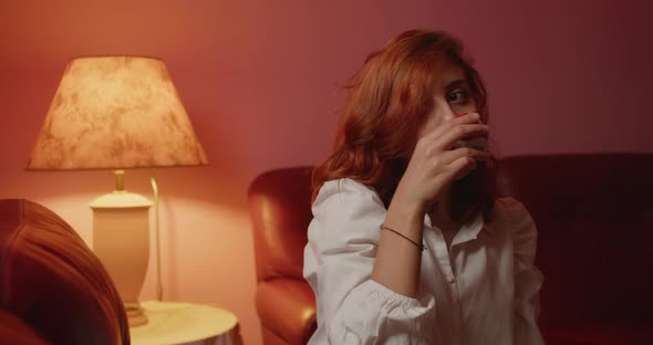 Italian girl with white shirt drinks coffee sitting on the sofa at home