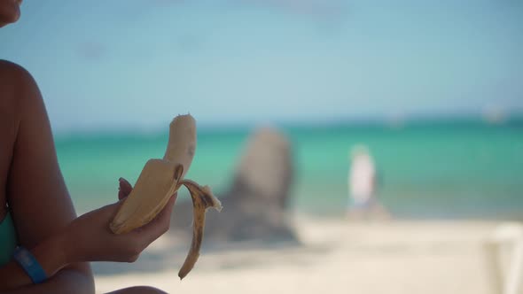 Girl Hand Holding Peeling And Eating Banana. Woman Eating Banana On Tropical Beach. Healthy Food.