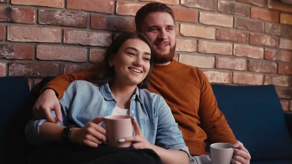 Young Hugging Couple Talking Holding Cups of Warm Drink and Watching Romantic Movie on TV Together