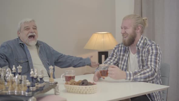 Two Cheerful Caucasian Men Laughing and Drinking Tea Indoors. Happy Young Son and Senior Father