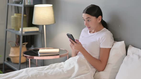 Success Latin Woman Celebrating on Smartphone in Bed