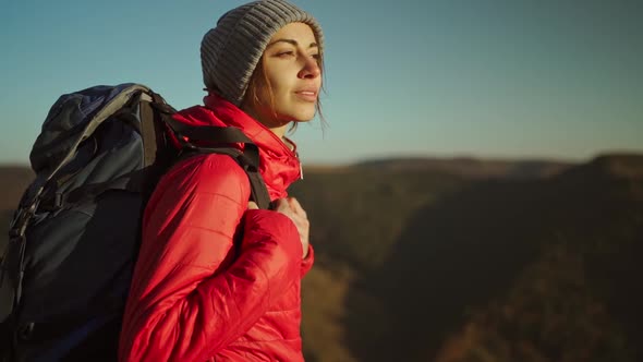 Slow Motion Portrait Woman Hiker in Bright Red Jacket with Backpack Stands on Mountain Top Against