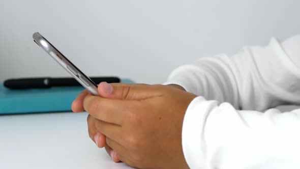 A child girl holds a phone in her hands, sits at the table, and does her homework.