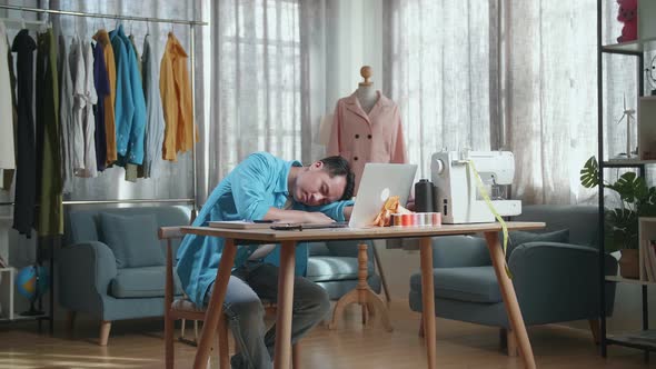 Asian Male Designer Working On A Laptop, Yawning, And Sleeping While Designing Clothes In The Studio