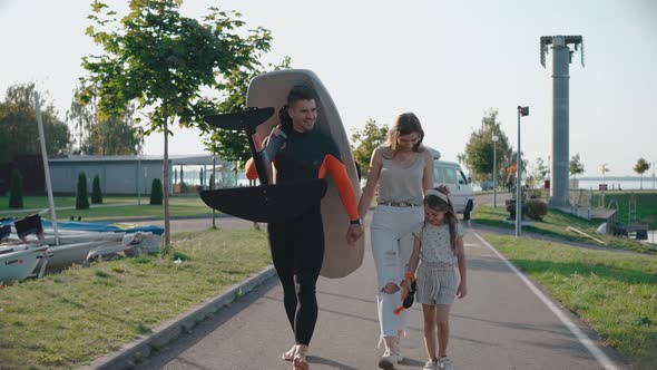 Man Walk with His Family Carrying a Surfboard to the Lake Shore on a Sunny Day