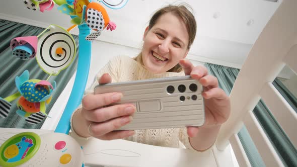 Happy Smiling Mother Making Video on Smartphone of Her Baby Lying in Cradle