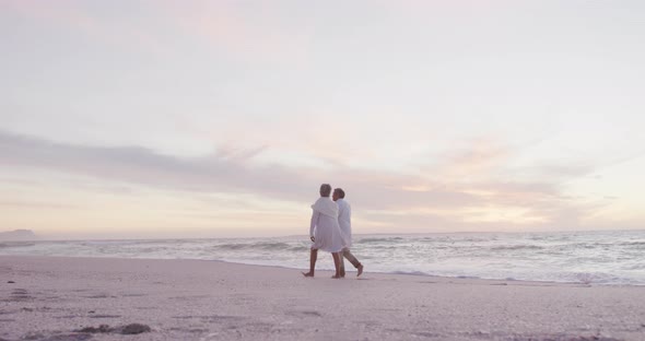 Back view of hispanic senior couple walking on beach at sunset