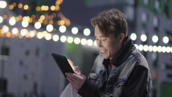 Young Man Uses a Tablet Typing and Talks on Video Near Modern Residential Area Conference Call