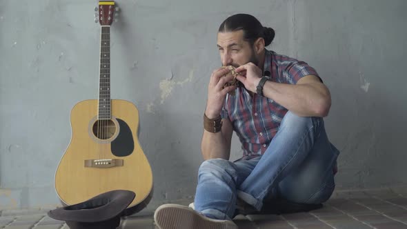 Wide Shot Front View of Young Frustrated Man Sitting in Underground Crossing with Guitar and Eating