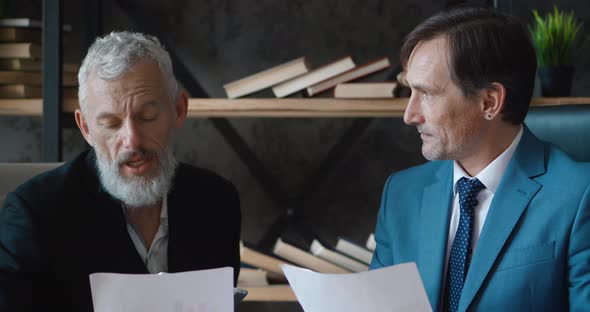 Two Mature Businessmen Discussing Business Growth in Modern Office While Sitting at Desk