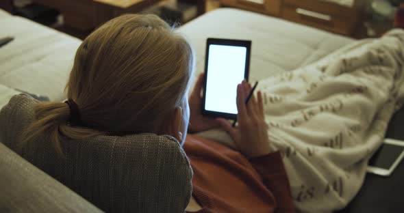 Young woman lies on the sofa under a cozy blanket and uses her tablet with a stylus