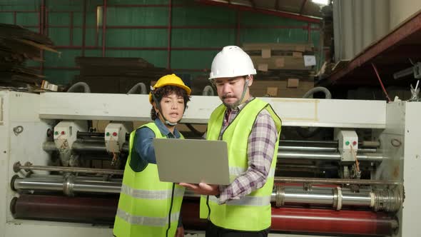 Two industrial workers use a laptop to control a paper manufacturing machine.