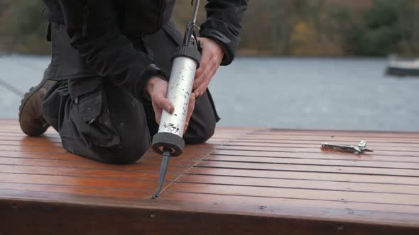 Young carpenter sealing roof planking of boat cabin with sealant gun in bleak weather.
