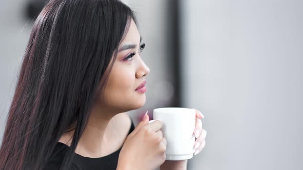 Charming Young Asian Smiling Girl Holding Big White Mug By Hands and Enjoying Hot Beverage