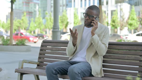 Angry African Man Talking on Phone While Sitting Outdoor on Bench