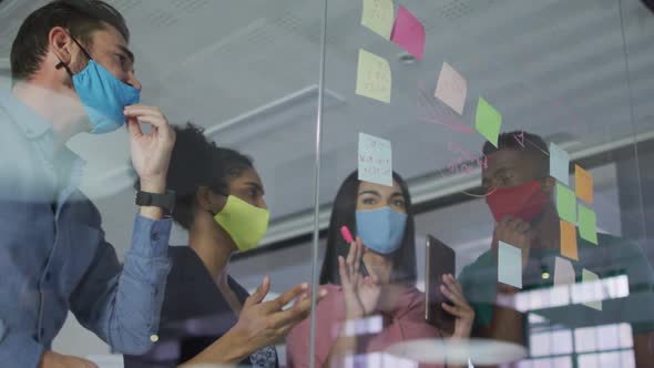 Diverse colleagues wearing face masks using memo notes on glass wall having a discussion