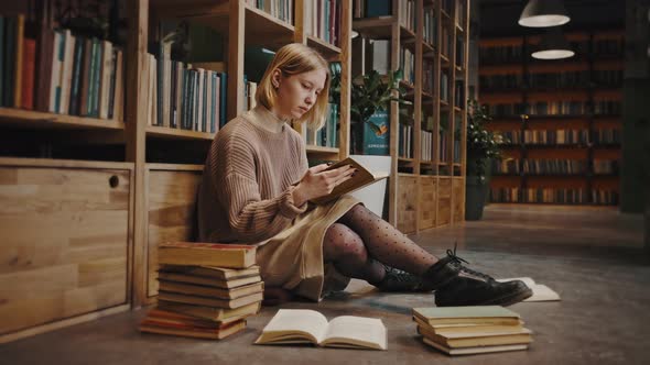 Young Woman Student Reading Book Sitting with Lots on Books on Floor at Library Leaning Bookshelf