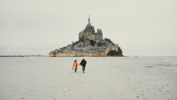 Drone Camera Follows Young Happy Couple Holding Hands Running Towards Epic Mont Saint Michel