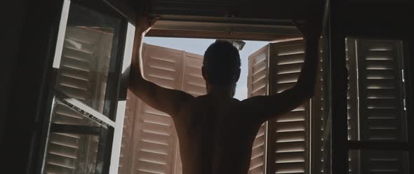 A shirtless man seen from behind leaning on the window shutter