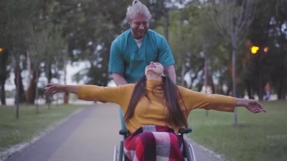 Portrait of Cheerful Paralyzed Woman with Stretched Hands Laughing As Male Nurse Running Rolling Her