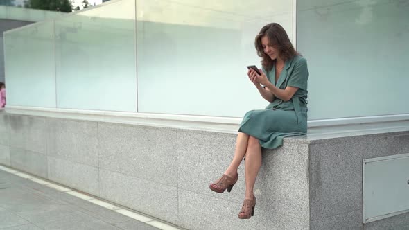 a Brunette in a Green Dress Sits on a Fence on a City Street and Holds a Phone in Her Hands