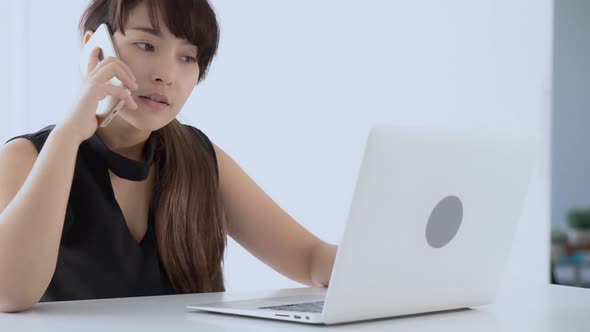 Young asian woman smiling working on laptop computer and talking customer with call phone at office.