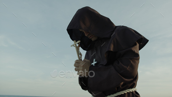 Religious Monk Prays With A Crucifix Of Jesus In His Hand Kneeling On The Sand