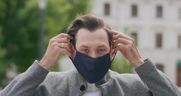 Young Man with Mustaches and a Beard Wears a Black Protective Mask Outdoor Buildings on Background