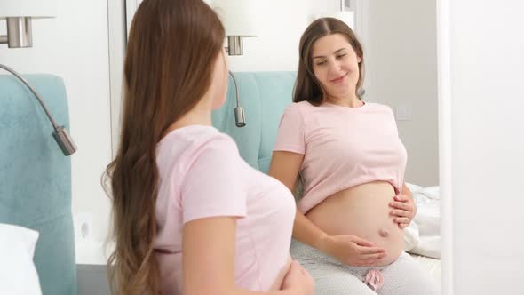 Beautiful Smiling Pregnant Woman Sitting on Bed and Looking in Her Reflection at Big Mirror