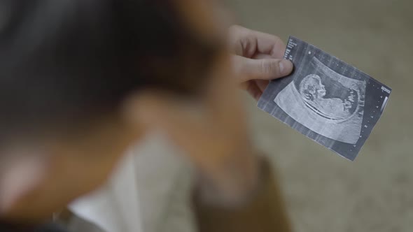 Unrecognizable Shocked Young Man Holding Ultrasound Picture in Hands and Touching Face