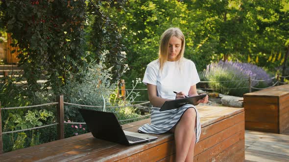 Young Woman is Sitting on a Bench of City Park and Drawing on a Digital Tablet Using Laptop