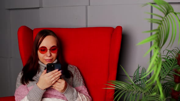 Woman in Stylish Sunglasses Sitting in Armchair with Smartphone