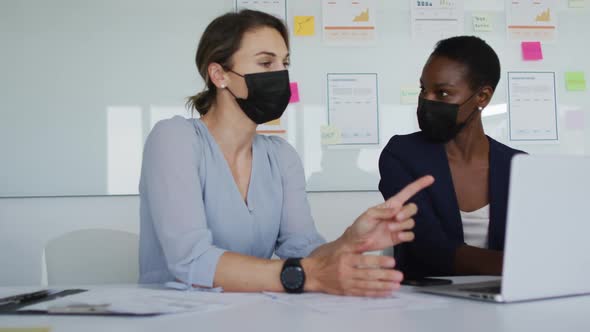 Two diverse female colleagues wearing face mask, sitting at desk, looking at laptop and talking