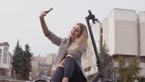 Young Stylish Blonde Girl in a Light Jacket and Jeans Sits on an Electric Scooter and Takes a Selfie