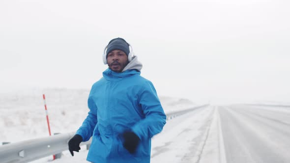 Black Man in Warm Clothes and Headphones Sprints Along Road Doing Outdoor Training in Winter Front