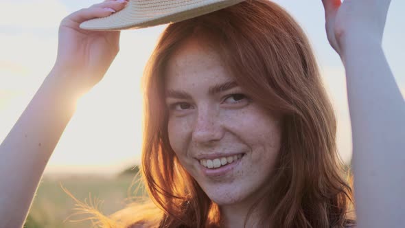 Cheerful Red Hair Girl with Freckles Puts on a Straw Hat on Her Head at Sunset