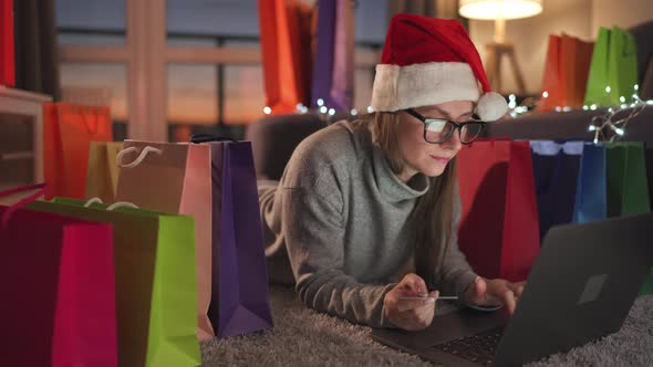 Happy Woman with Glasses Wearing a Santa Claus Hat Is Lying on the Carpet and Makes an Online
