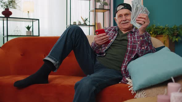 Smiling Senior Man Holding Money and Smartphone Excited About Mobile App Sport Bet Bid Win at Home