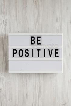 'Be positive' words on a lightbox on a white wooden background, top view.