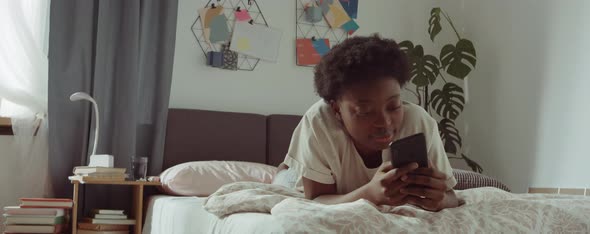 Cheerful Black Woman Lying on Bed and Using Smartphone