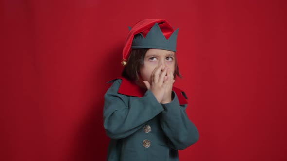 Baby Boy in a Green Suit and a Dwarf Hat is Indulging on Red Background for Christmas