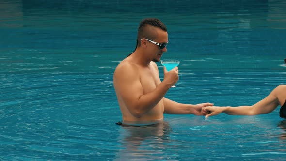 The Loving Couple Hugs and Kisses Drinking Blue Cocktail Alcohol Liquor in Swimming Pool at Hotel