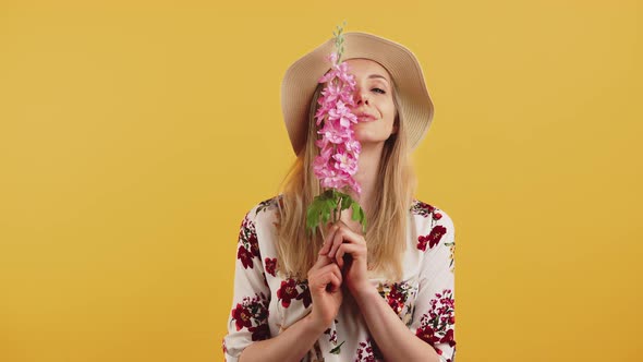 Young Caucasian Blonde Woman Wearing Summer Clothes Holding Pink Flower with Both Hands Smelling It