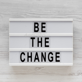 'Be the change' words on a lightbox on a white wooden background, top view.