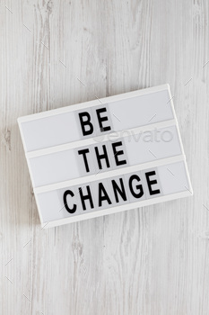 'Be the change' words on a lightbox on a white wooden background, top view.