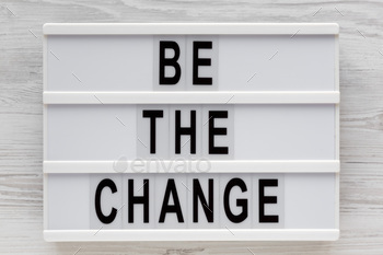 'Be the change' words on a lightbox on a white wooden surface, top view.