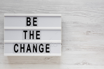'Be the change' words on a lightbox on a white wooden surface, top view.